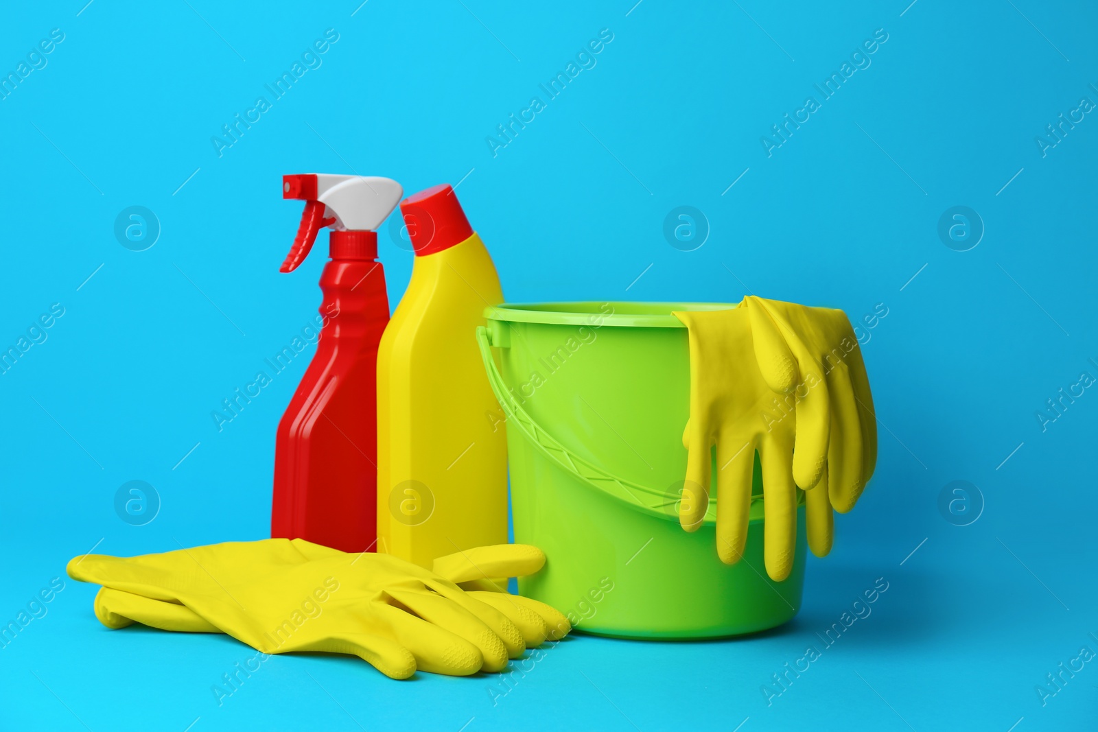 Photo of Green bucket, gloves and bottles of detergents on light blue background. Cleaning supplies