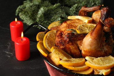 Photo of Baked chicken with orange slices and burning candles on black table, closeup