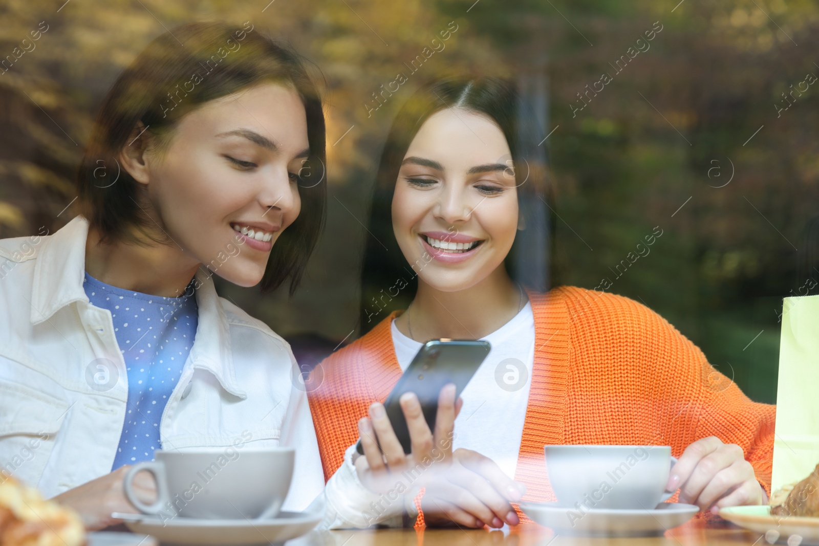 Photo of Special Promotion. Happy young women using smartphone at table in cafe, view from outdoors