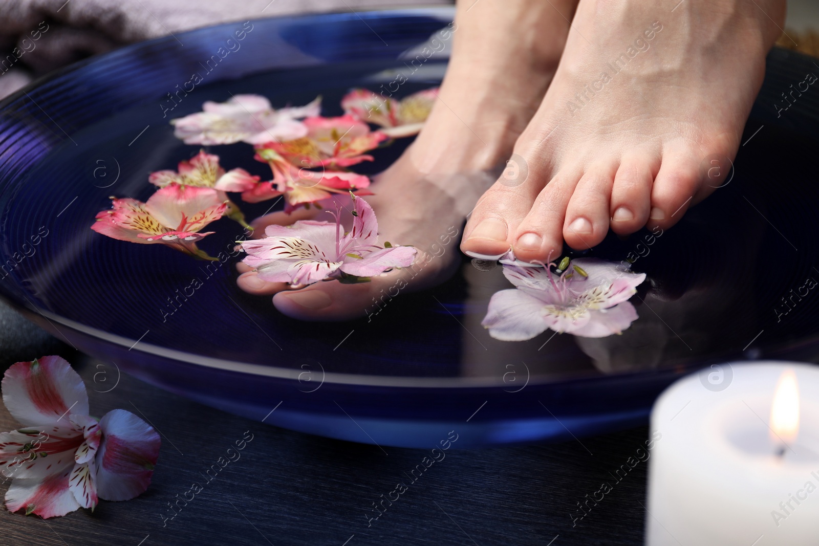 Photo of Woman soaking her feet in bowl with water and flowers on wooden floor, closeup. Spa treatment