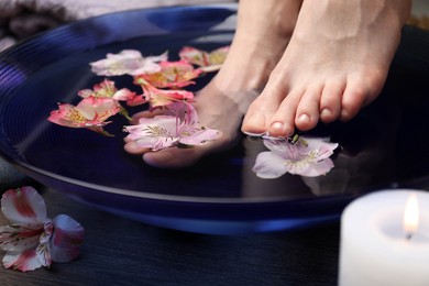 Photo of Woman soaking her feet in bowl with water and flowers on wooden floor, closeup. Spa treatment