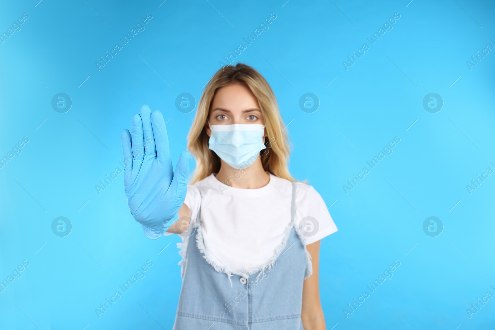 Photo of Woman in protective mask showing stop gesture on light blue background. Prevent spreading of COVID‑19