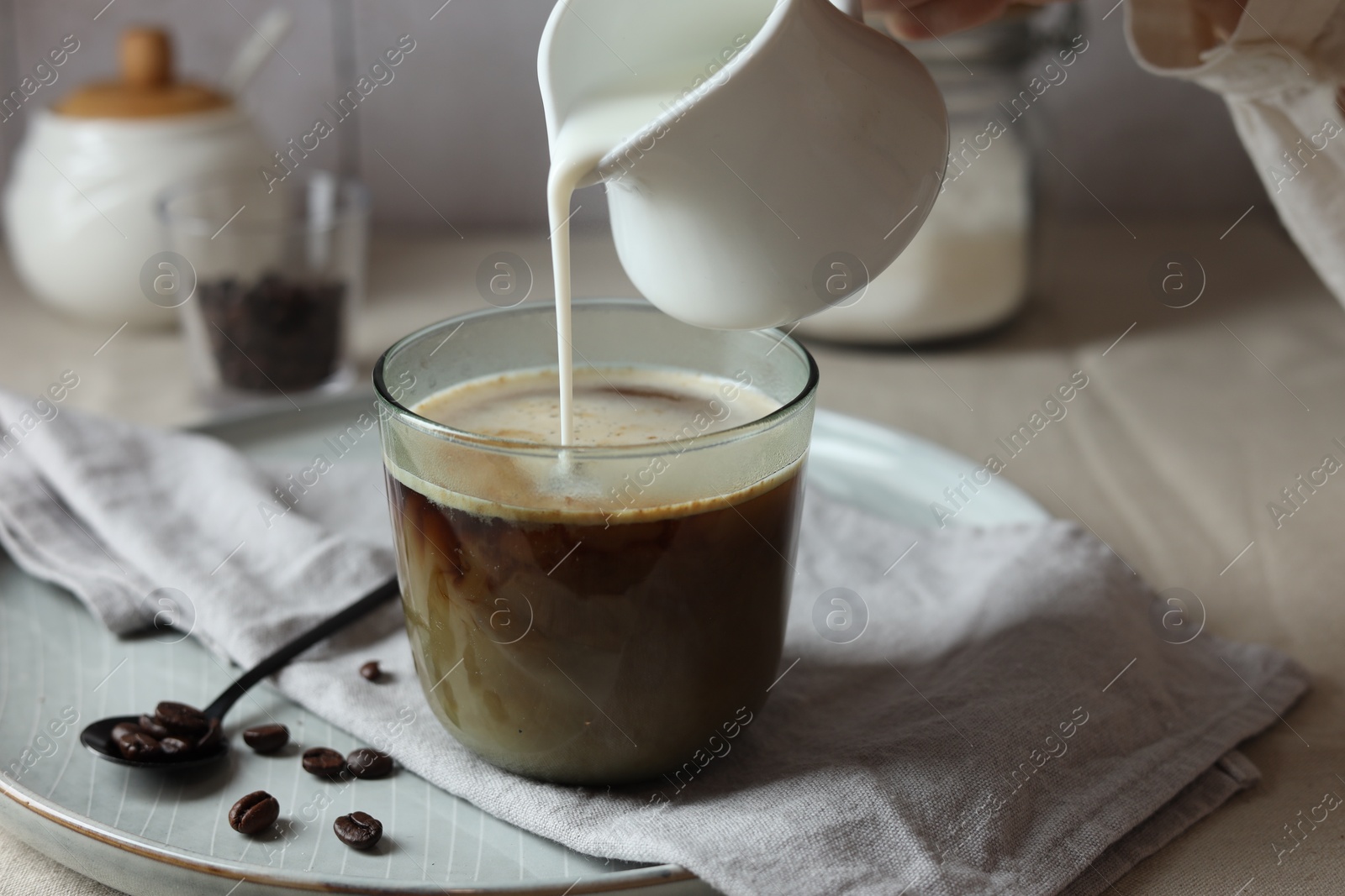 Photo of Woman pouring milk into cup with coffee at light table, closeup