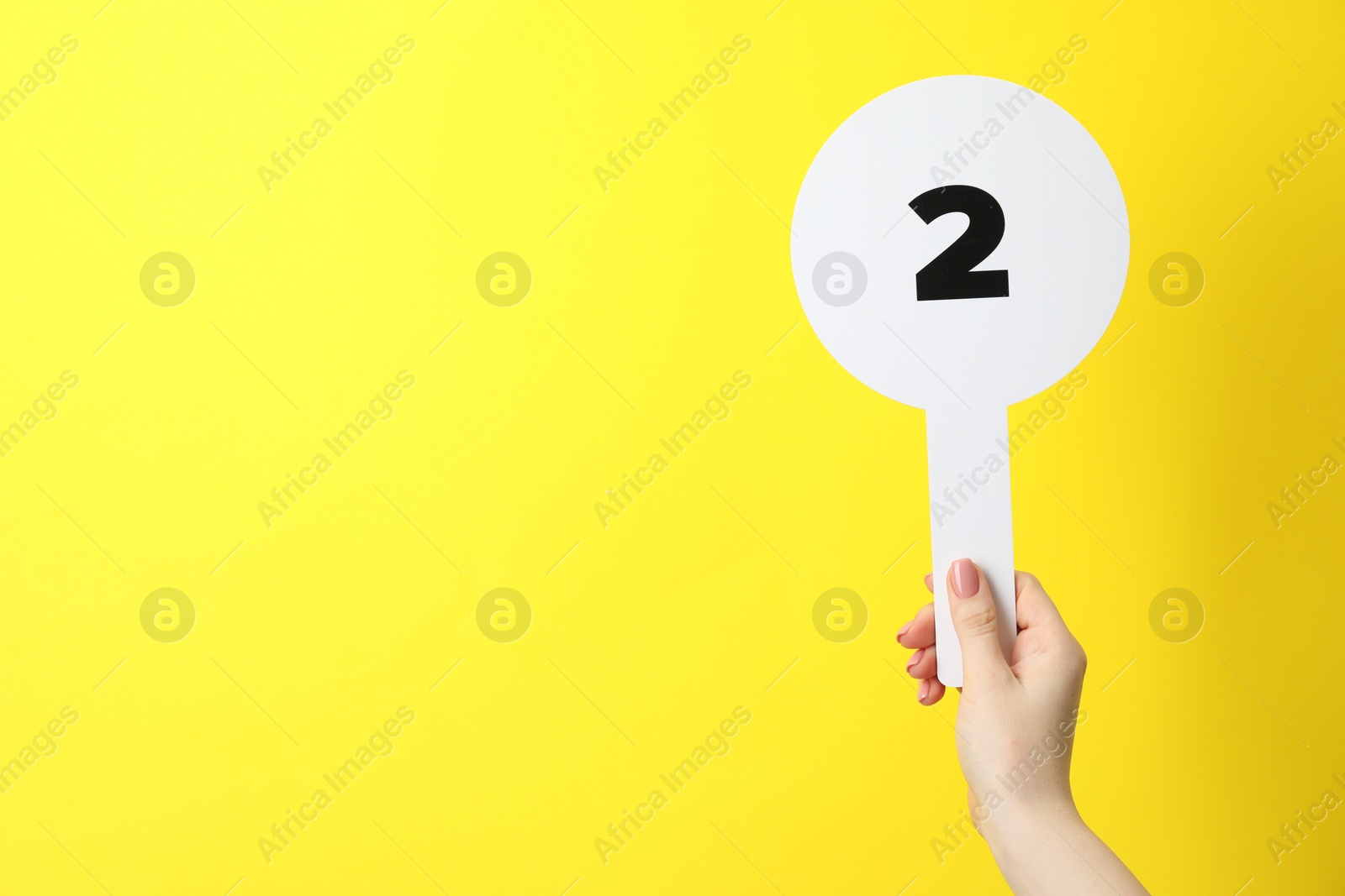 Photo of Woman holding auction paddle with number 2 on yellow background, closeup. Space for text