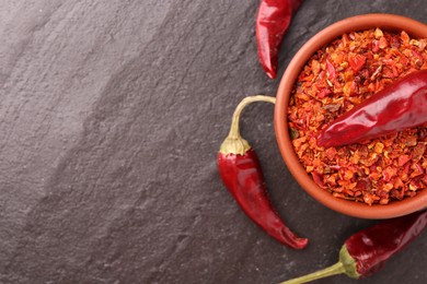 Photo of Chili pepper flakes in bowl and pods on dark textured table, flat lay. Space for text