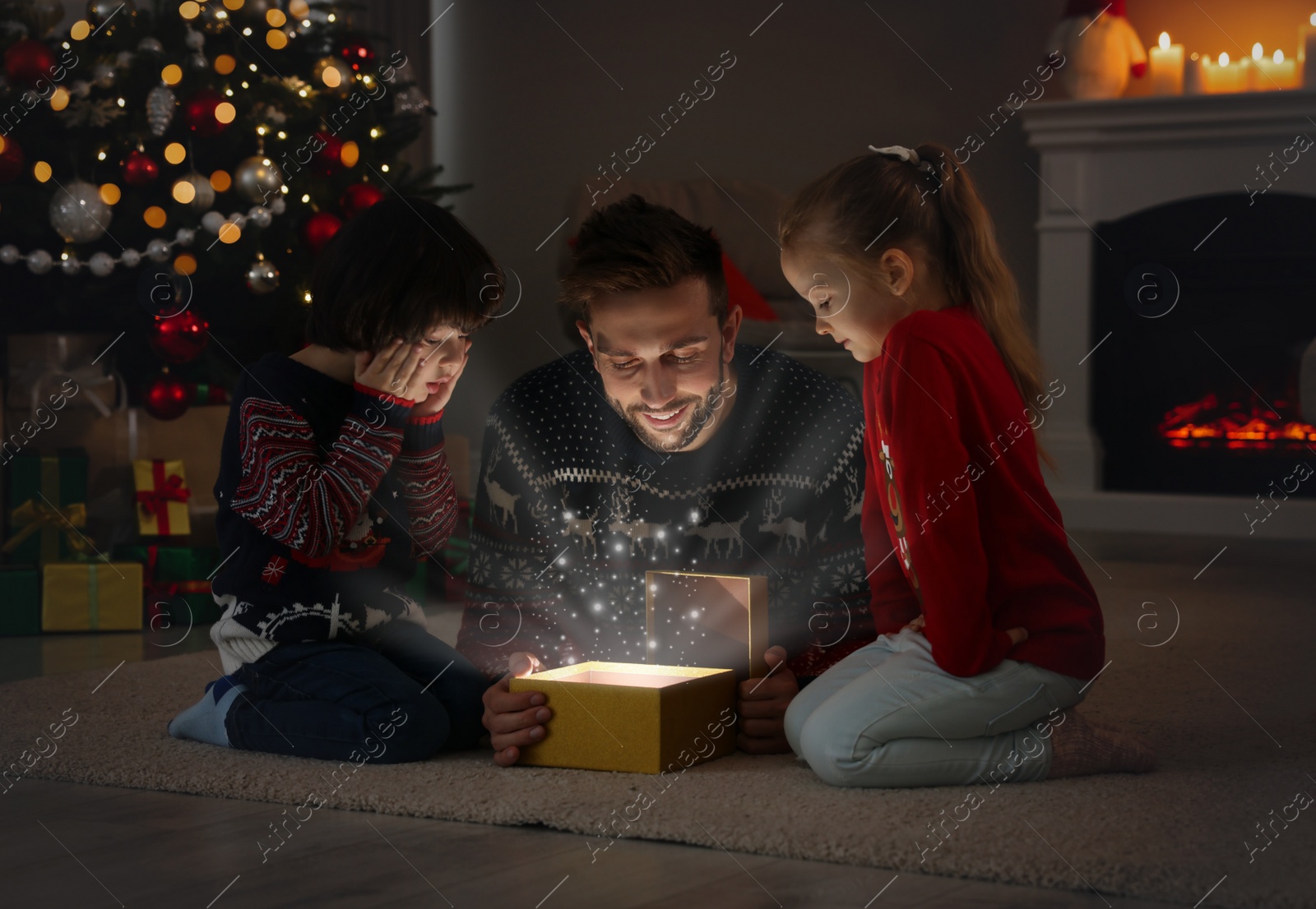 Photo of Father and his cute children opening gift box with magical light on floor at home. Christmas celebration