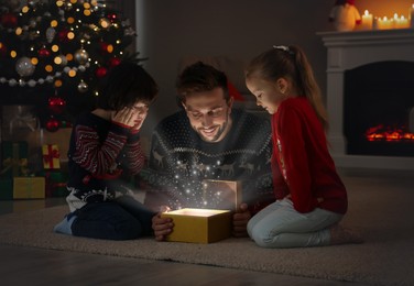 Photo of Father and his cute children opening gift box with magical light on floor at home. Christmas celebration