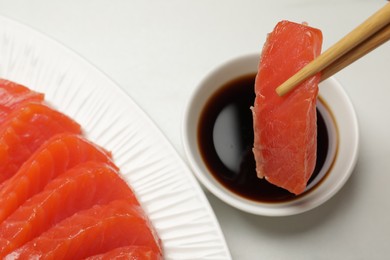 Dipping tasty salmon slice into soy sauce with chopsticks at white table, above view. Delicious sashimi dish