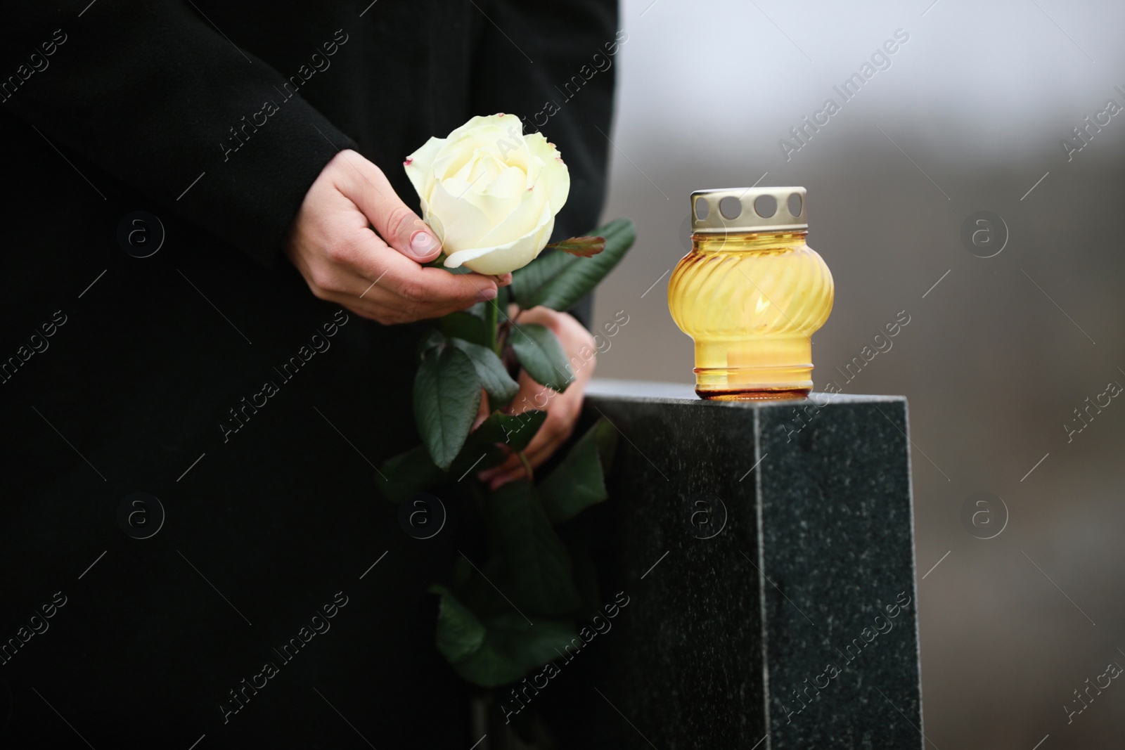 Photo of Woman holding white rose near black granite tombstone with candle outdoors, closeup. Funeral ceremony