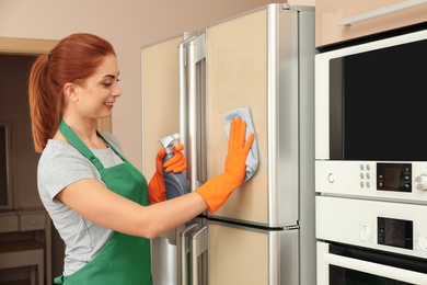 Woman in protective gloves cleaning refrigerator with rag indoors