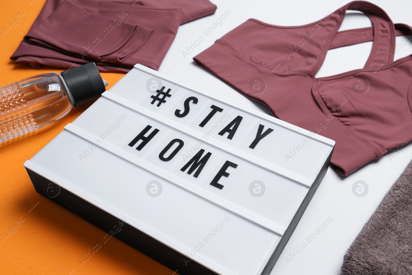 Photo of Stylish sportswear, yoga mat and light box with hashtag Stay at Home on white background, closeup. Self isolation during COVID‑19 pandemic
