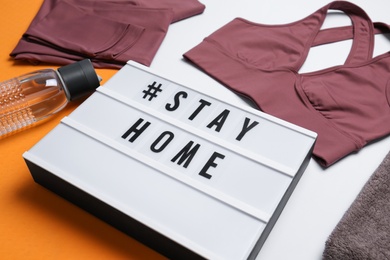 Stylish sportswear, yoga mat and light box with hashtag Stay at Home on white background, closeup. Self isolation during COVID‑19 pandemic