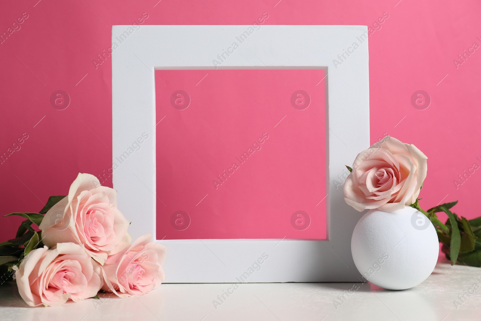 Photo of Stylish presentation for product. Frame, beautiful roses and ball on light table against pink background, space for text