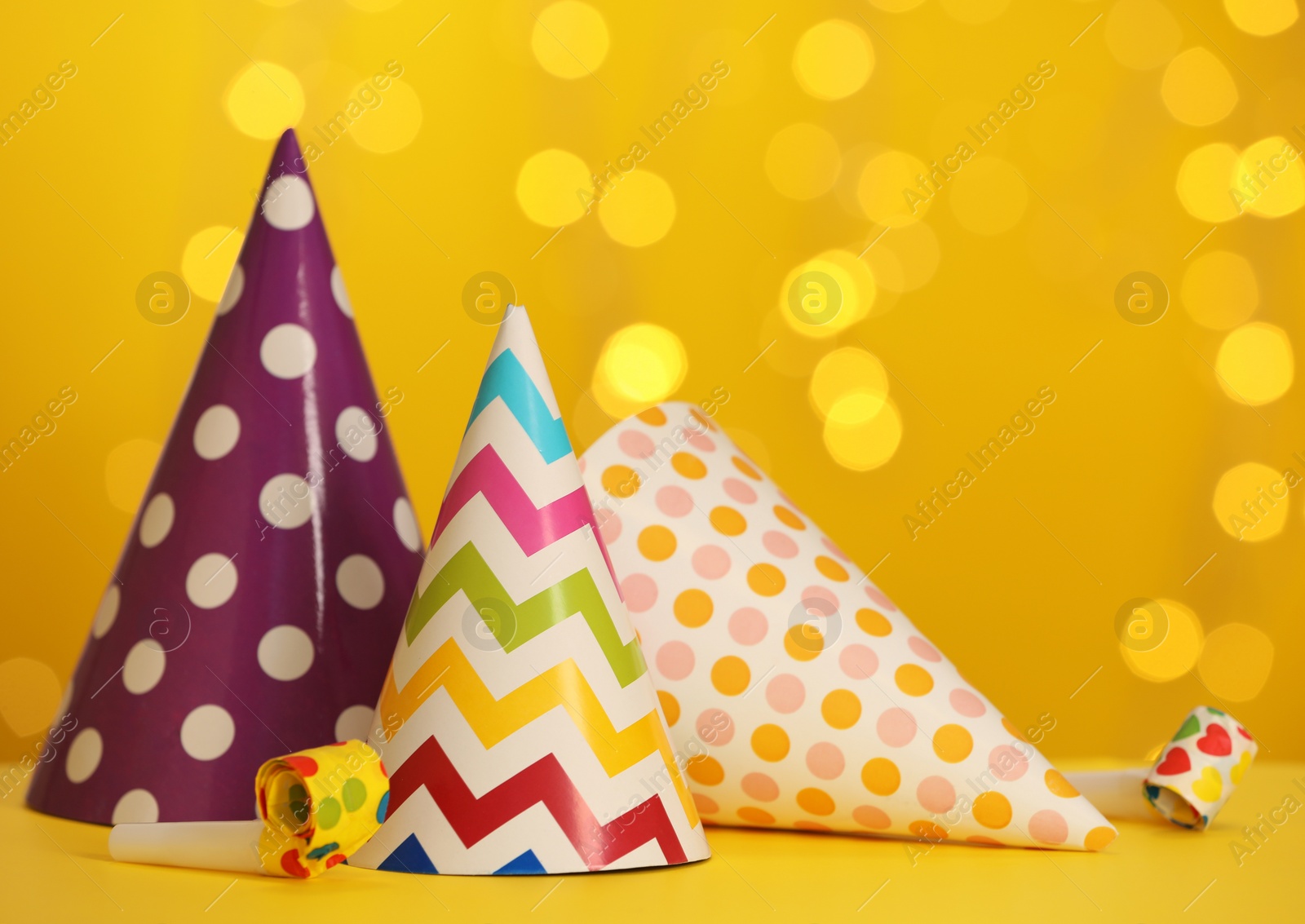 Photo of Beautiful party hats and blowers on yellow table against blurred festive lights. Space for text