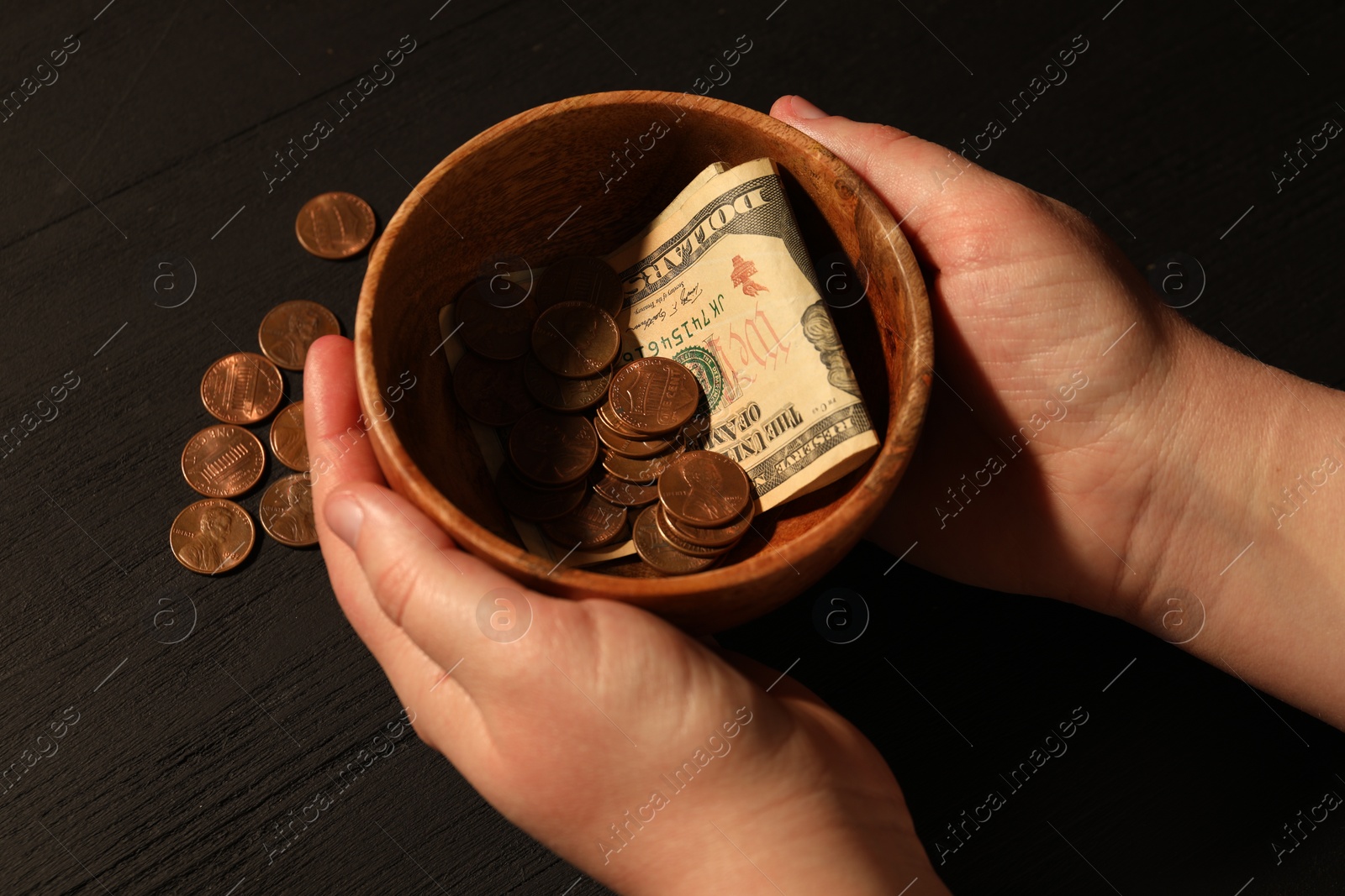 Photo of Donate and give concept. Woman holding bowl with coins and dollar banknotes at black wooden table, closeup