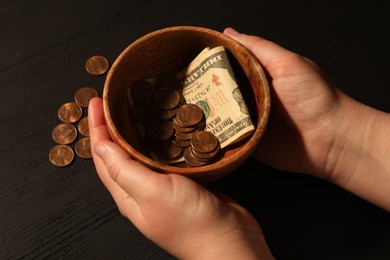 Photo of Donate and give concept. Woman holding bowl with coins and dollar banknotes at black wooden table, closeup