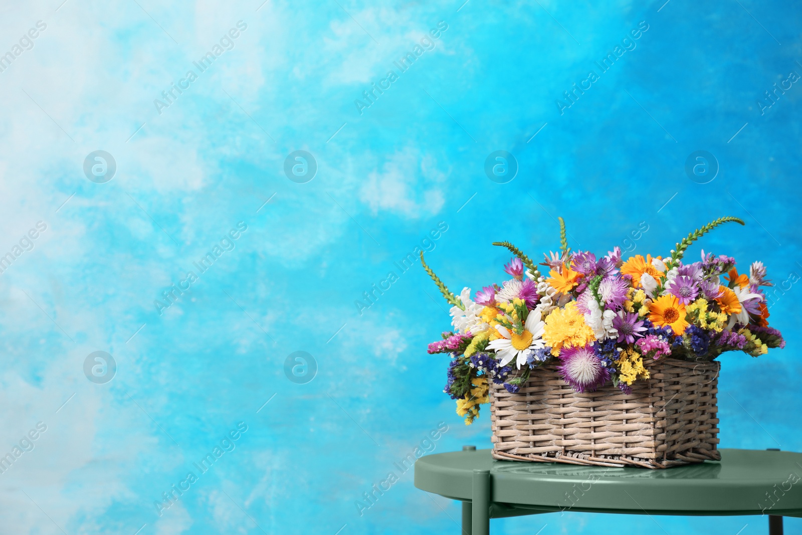 Photo of Wicker basket with beautiful wild flowers on table against color background