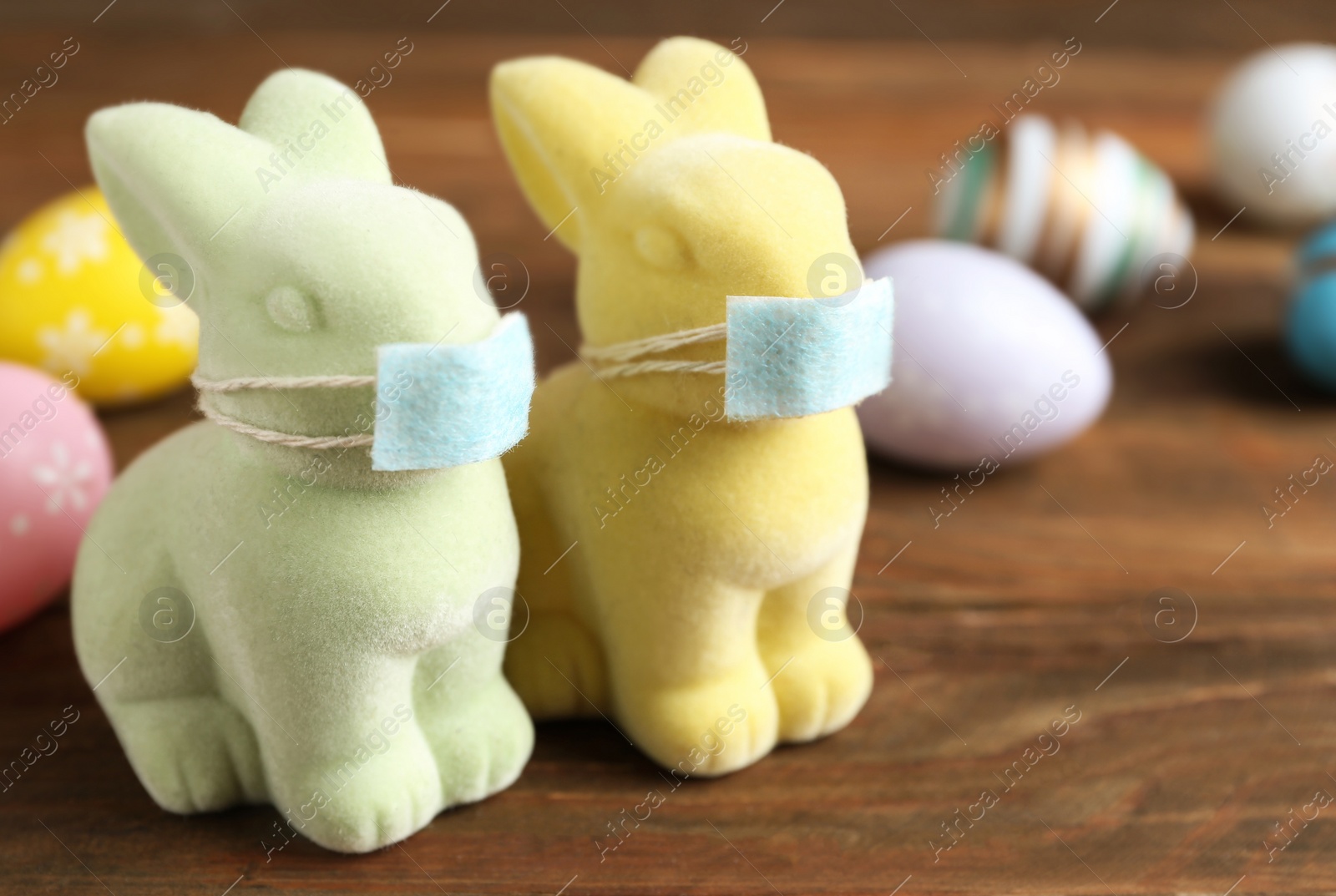 Photo of Cute bunny figures in protective masks near dyed eggs on wooden table. Easter holiday during COVID-19 quarantine