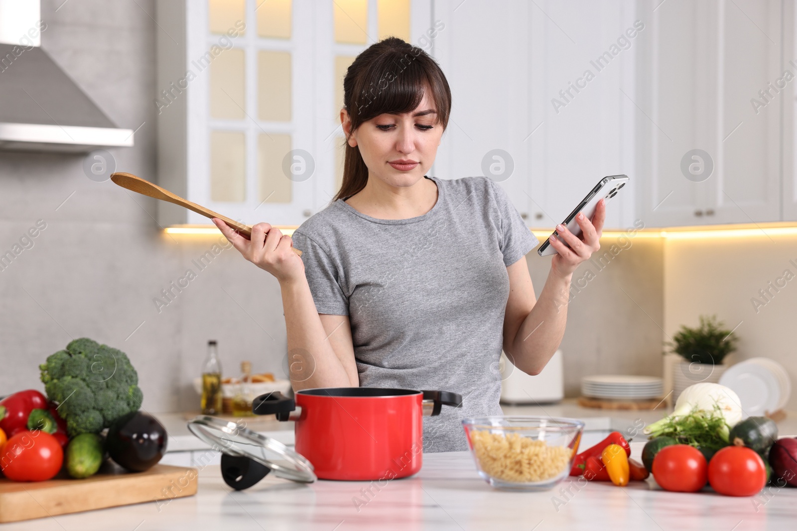 Photo of Young housewife with spoon using smartphone while cooking at white marble table in kitchen