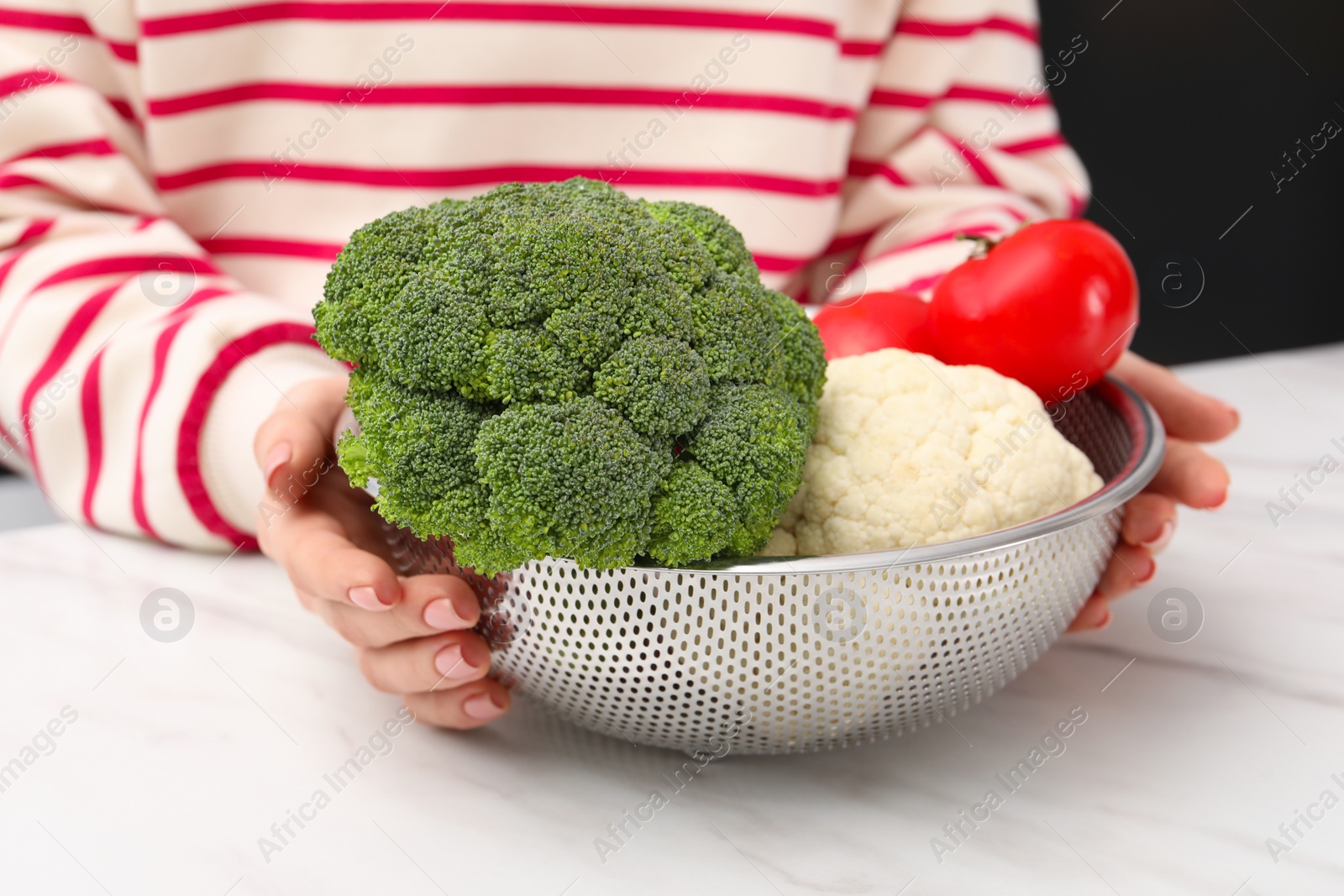 Photo of Woman holding colander with fresh vegetables at white marble table, closeup