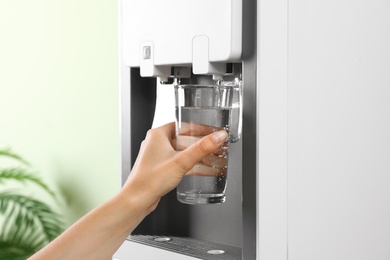 Photo of Woman filling glass from water cooler indoors, closeup. Refreshing drink