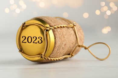 Cork of sparkling wine and muselet cap with engraving 2023 on table against blurred festive lights, closeup. Bokeh effect