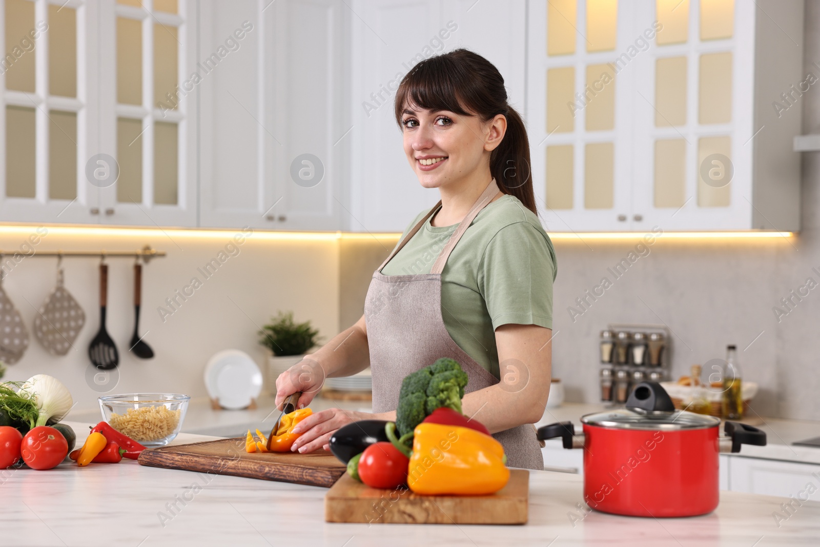 Photo of Happy young housewife cutting bell pepper at white marble table in kitchen