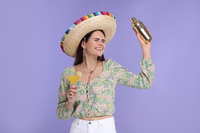 Young woman in Mexican sombrero hat with cocktail and shaker on violet background