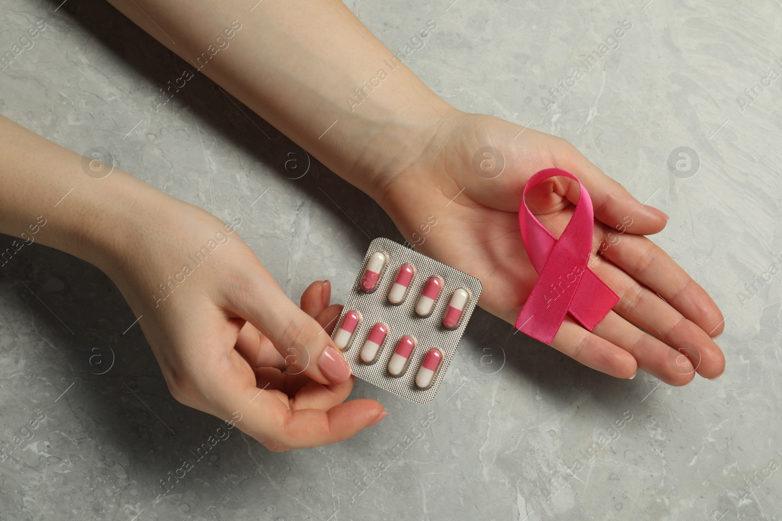 Photo of Woman with pink ribbon and pills at grey marble table, top view. Breast cancer awareness