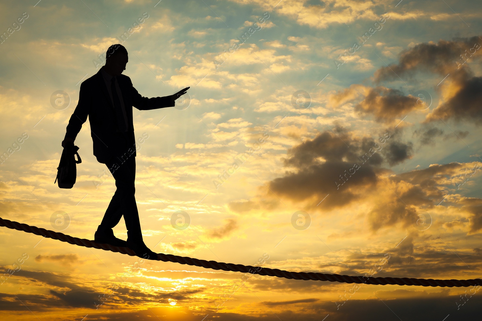 Image of Risks and challenges of owning business. Silhouette of man balancing on rope in sunset sky