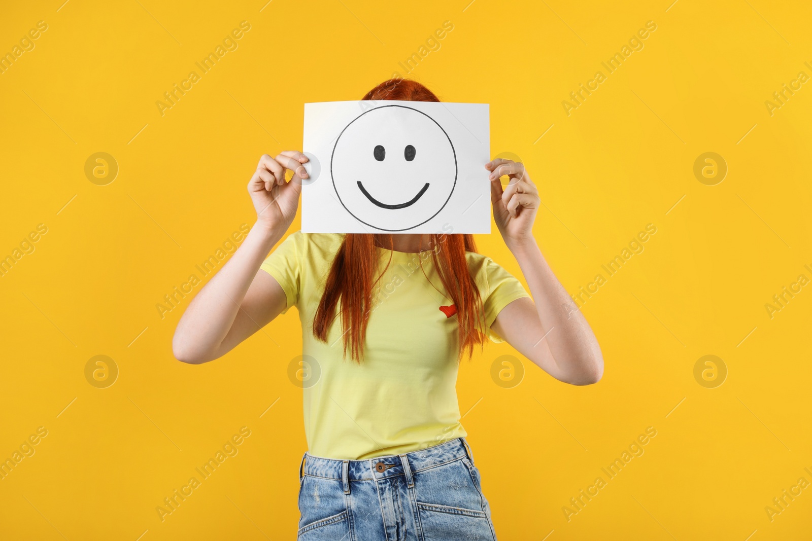 Photo of Woman hiding behind sheet of paper with happy face on yellow background