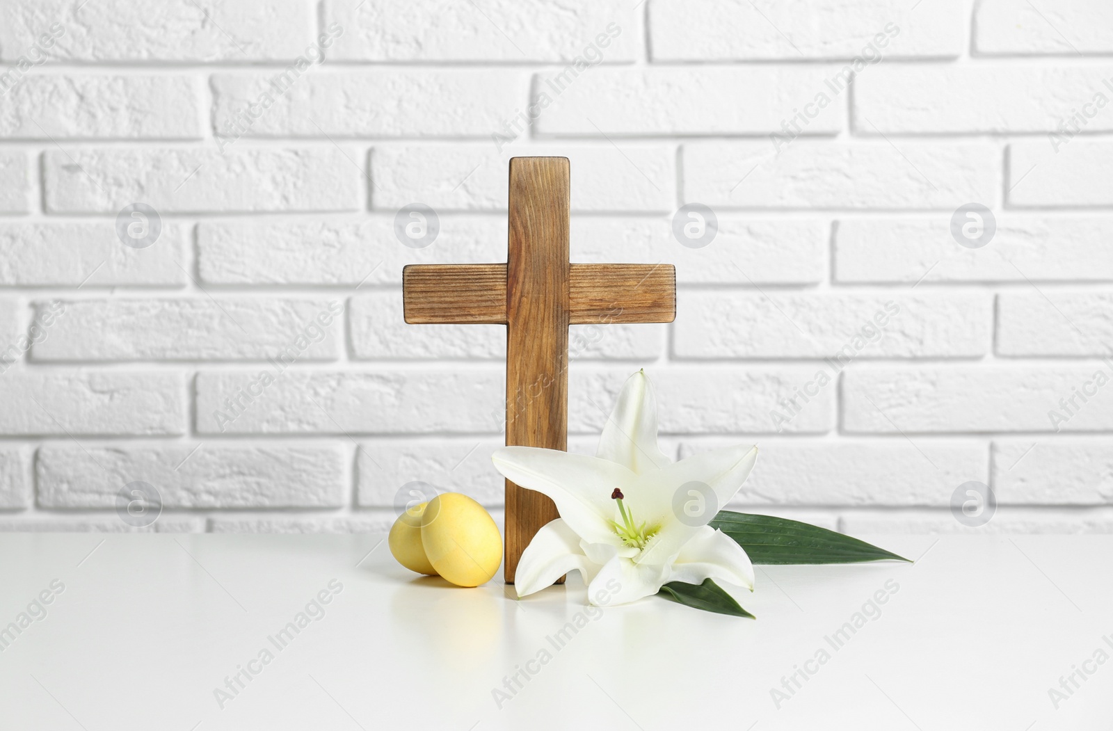 Photo of Wooden cross, Easter eggs and blossom lily on table against brick wall