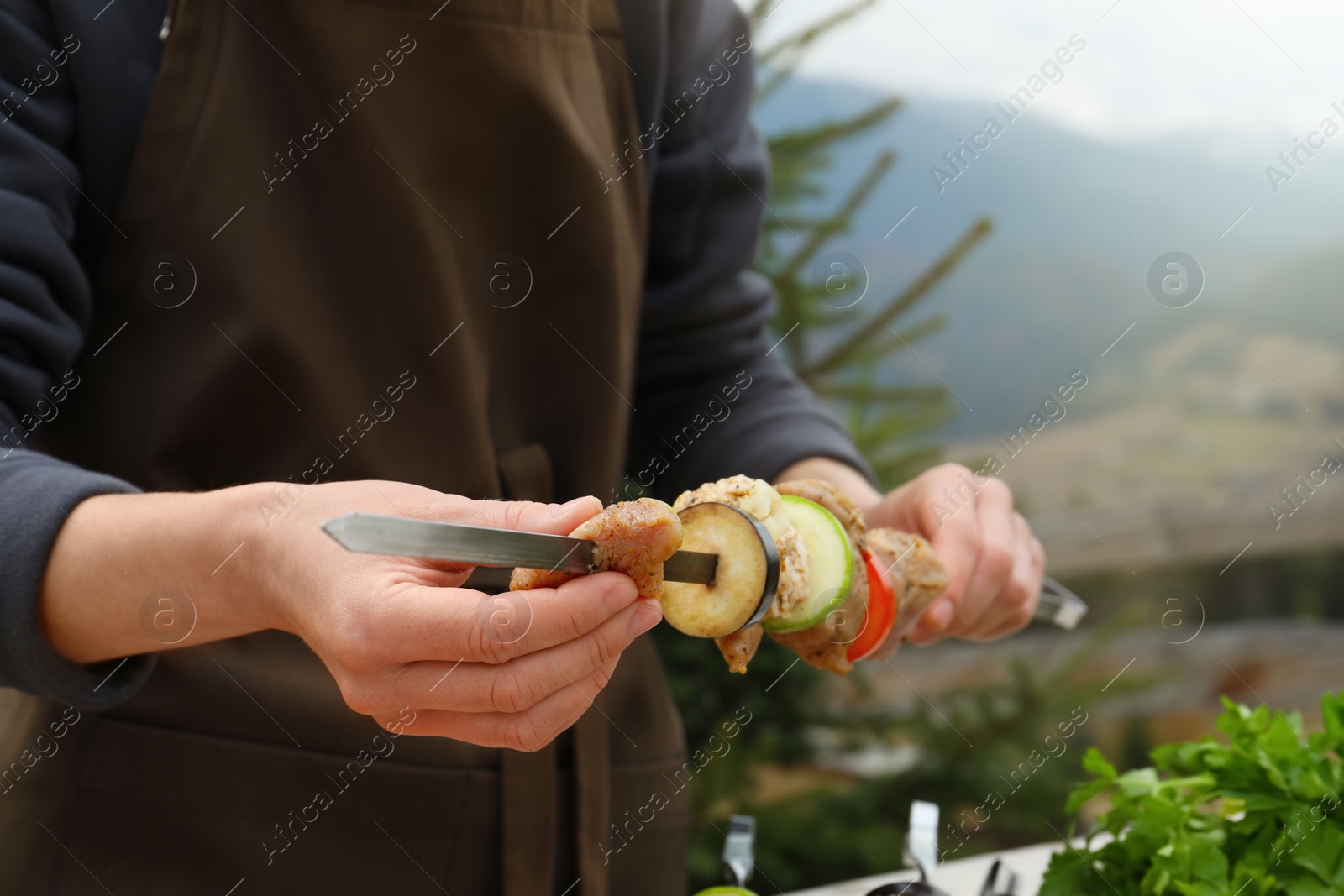 Photo of Woman stringing marinated meat and vegetables on skewer against mountain landscape, closeup