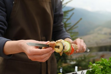 Photo of Woman stringing marinated meat and vegetables on skewer against mountain landscape, closeup