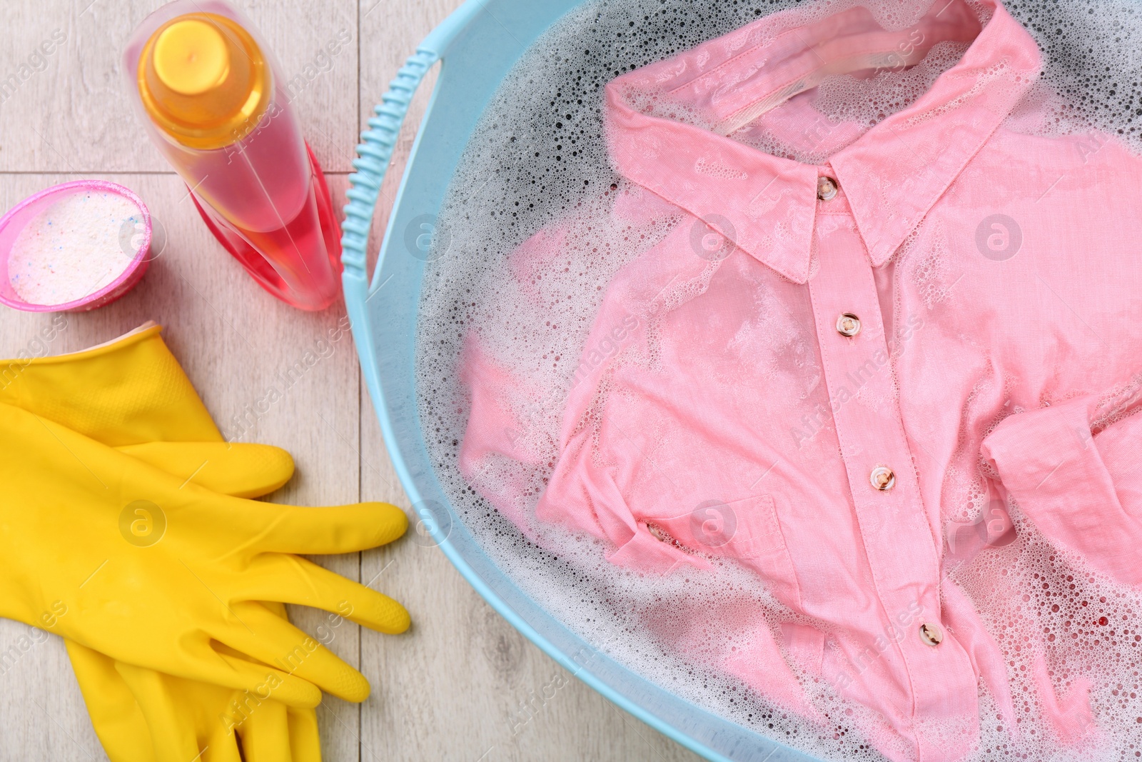Photo of Pink shirt in basin, powder, gloves and bottle of detergent on floor, flat lay. Hand washing laundry