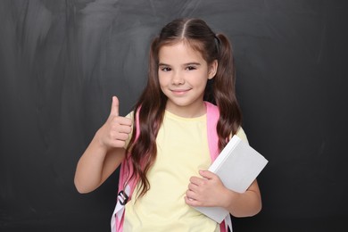 Photo of Cute schoolgirl with book showing thumbs up near chalkboard