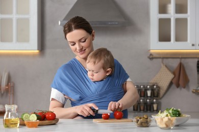Mother cutting tomatoes while holding her child in sling (baby carrier) in kitchen