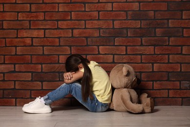 Photo of Child abuse. Upset little girl with teddy bear sitting on floor near brick wall indoors