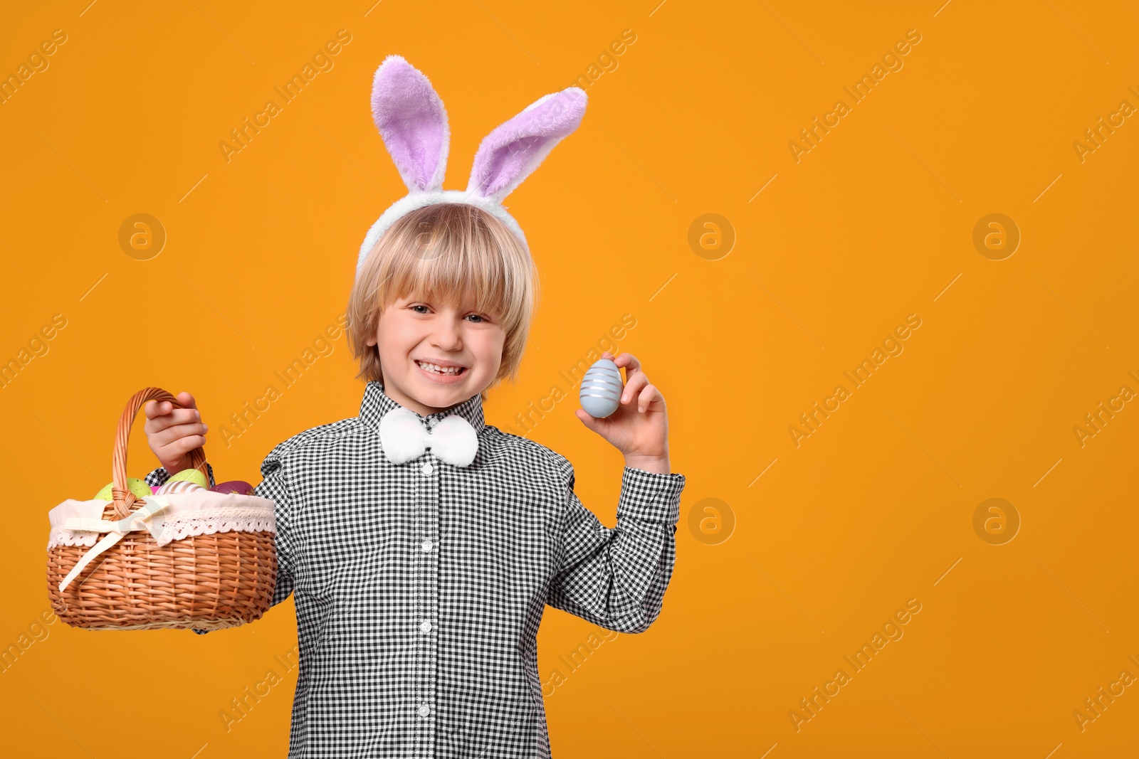 Photo of Happy boy in bunny ears headband holding wicker basket with painted Easter eggs on orange background. Space for text