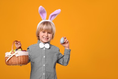 Photo of Happy boy in bunny ears headband holding wicker basket with painted Easter eggs on orange background. Space for text