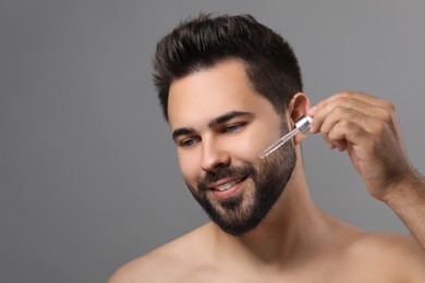 Handsome man applying cosmetic serum onto face on grey background, space for text