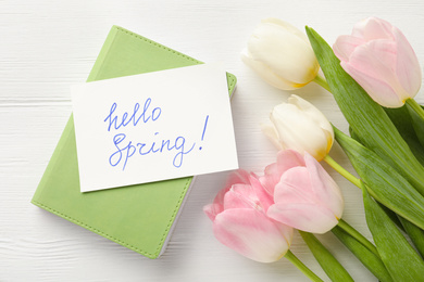 Card with words HELLO SPRING, notebook and tulips on white wooden table, flat lay