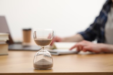Photo of Hourglass with flowing sand on desk. Man taking notes indoors, selective focus