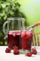 Refreshing hibiscus tea with ice cubes and roselle flowers on white wooden table against blurred green background