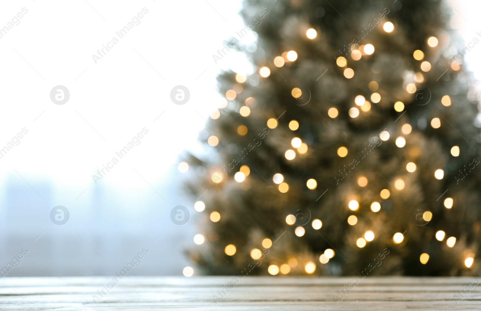 Photo of Blurred view of beautiful Christmas tree with yellow lights near window indoors, focus on wooden table. Space for text