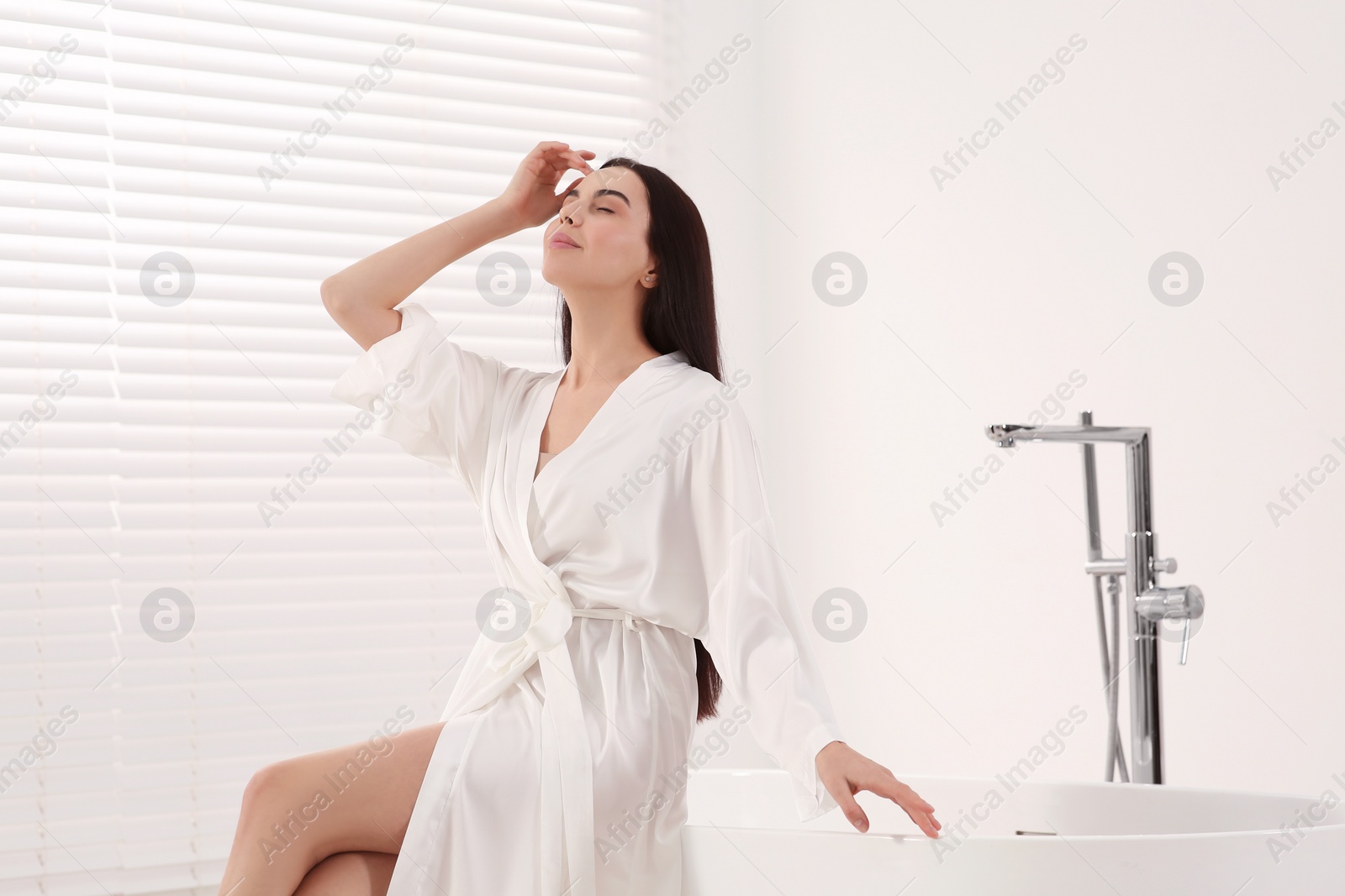 Photo of Beautiful happy woman in stylish bathrobe sitting on tub in bathroom