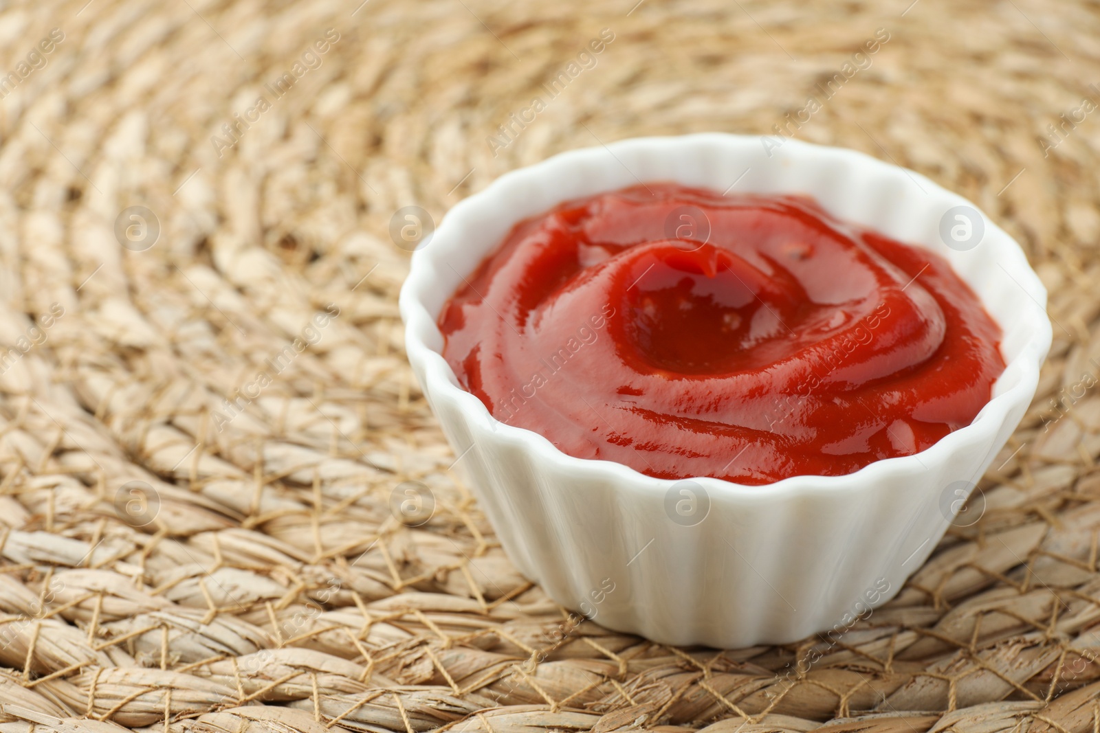 Photo of Bowl of tasty ketchup on wicker mat, closeup. Space for text