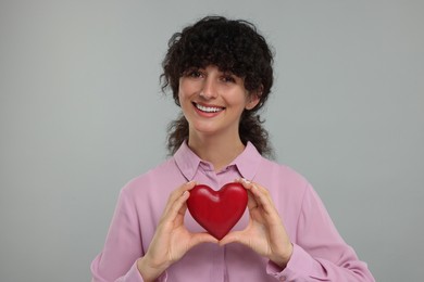 Happy young woman holding decorative red heart on light grey background