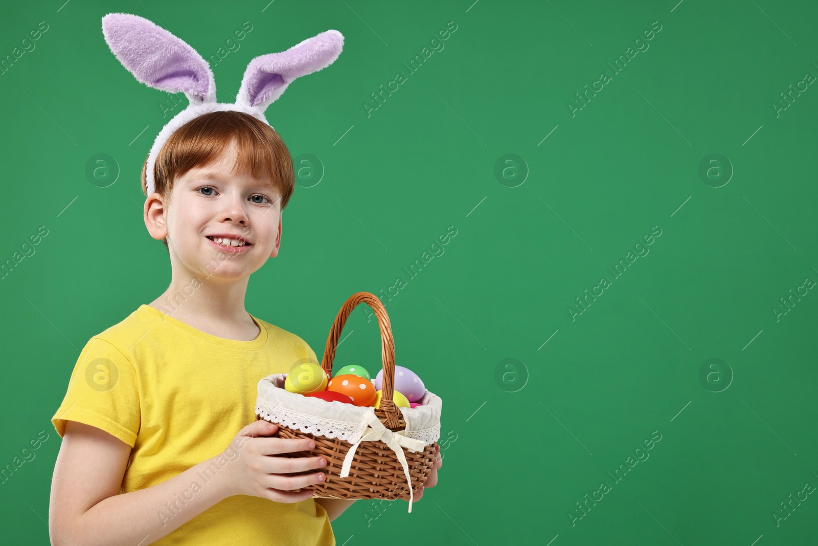Photo of Easter celebration. Cute little boy with bunny ears and wicker basket full of painted eggs on green background. Space for text