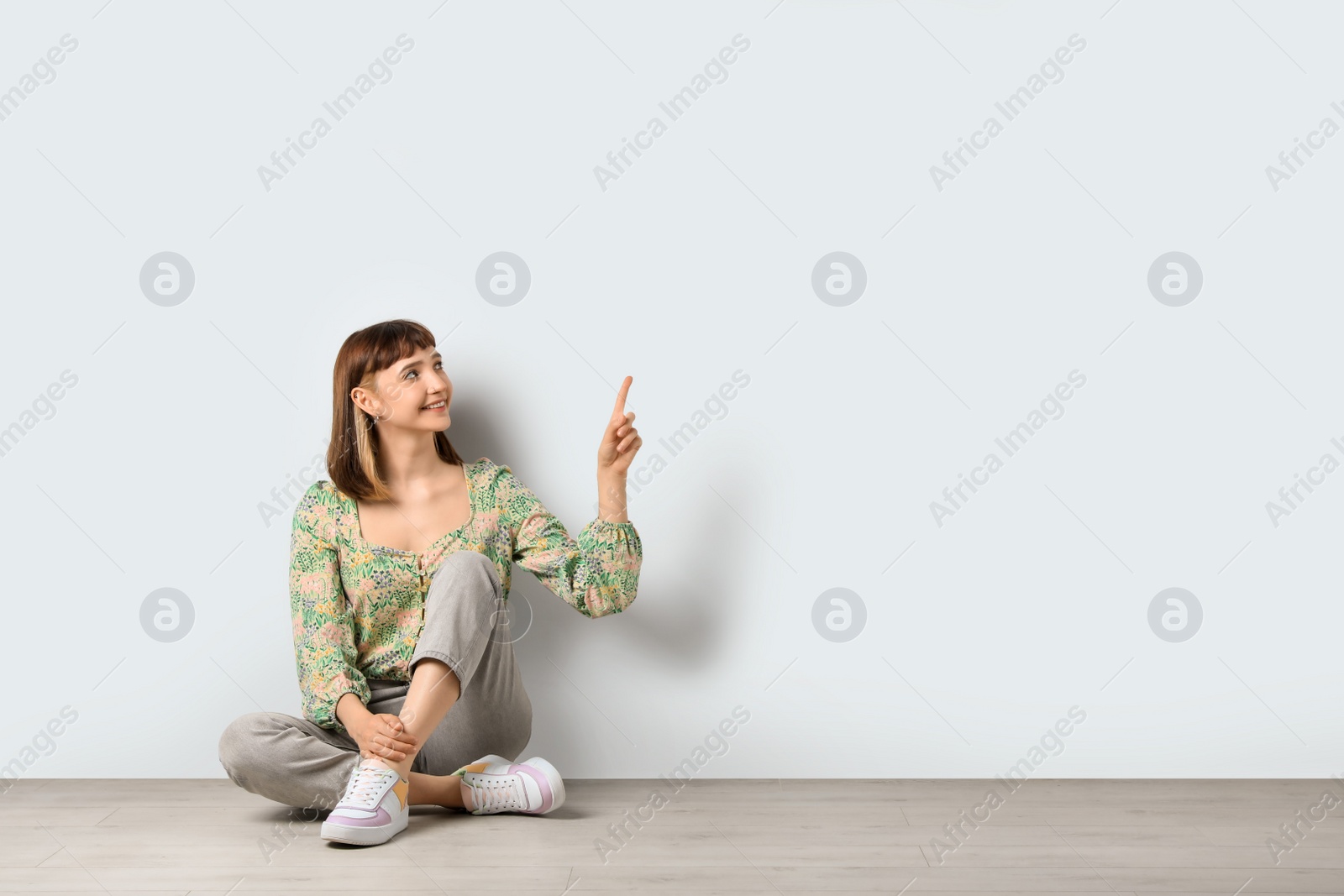 Photo of Beautiful young girl sitting on floor and pointing at something against white background. Space for text